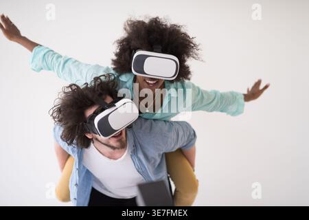 Couple noir et caucasien portant des casques VR ayant un plaisir piggyback équitation et expérimentant la réalité virtuelle sur fond blanc en studio Banque D'Images