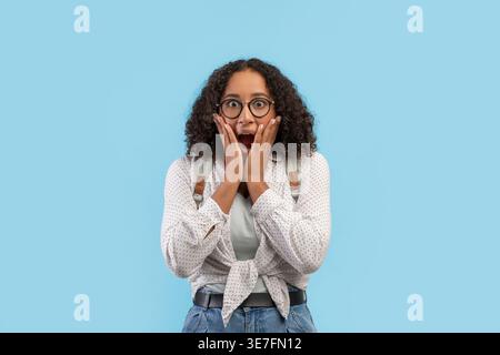 Adolescente noire portant des lunettes avec une expression choquée debout avec les mains sur le visage sur fond bleu en studio Banque D'Images