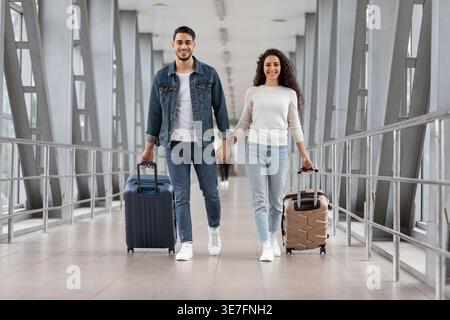 Couple du moyen-Orient regardant loin tout en se tenant la main marchant avec une valise à l'aéroport Banque D'Images