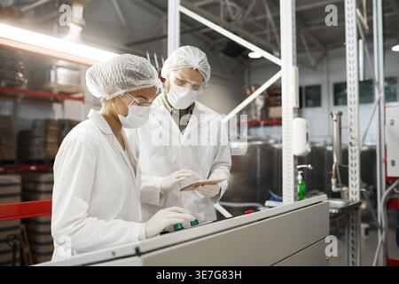 Femmes mixtes et hommes du caucase portant des costumes de protection et des masques actionnant la machine tout en se tenant debout dans l'usine chimique Banque D'Images