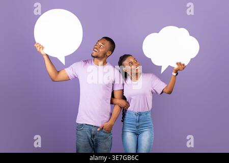 Sourire couple noir tenant et regardant des bulles de parole tout en se tenant debout sur fond violet dans le studio Banque D'Images