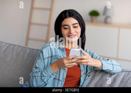 Femme arabe utilisant un smartphone tout en étant assis sur le canapé à la maison Banque D'Images