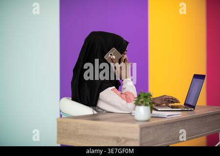 Femme d'affaires musulmane noire portant le hijab ayant un appel téléphonique et travaillant sur un ordinateur portable tout en étant assise à table contre un mur coloré dans le bureau Banque D'Images