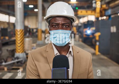 Ingénieur masculin noir portant un casque de sécurité et un masque facial tenant le microphone debout dans l'entrepôt Banque D'Images