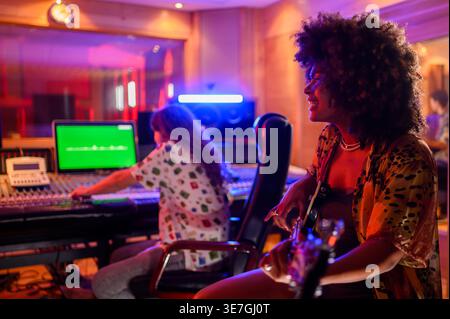 Musicienne hispanique souriante regardant loin assis et jouant de la guitare près d'un producteur de musique masculin caucasien pendant la session d'enregistrement en studio Banque D'Images