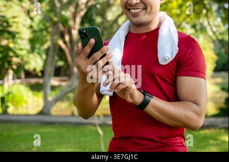 Image recadrée de l'homme thaïlandais dans des vêtements de fitness à l'aide d'un smartphone tout en se tenant debout avec une serviette blanche autour de son cou dans le parc le jour ensoleillé Banque D'Images