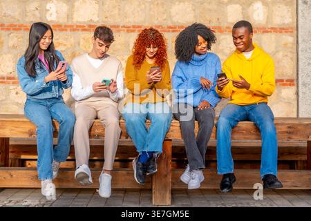 Souriant un groupe d'amis diversifié à l'aide de smartphones tout en étant assis ensemble sur un banc à l'extérieur pendant la journée Banque D'Images