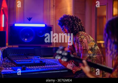 Musicienne hispanique sérieuse avec afro regardant loin assis à côté d'une console de mixage avec lumière néon écoutant l'enregistrement de la lecture de guitare... Banque D'Images