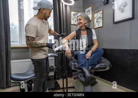 Tatoueur masculin asiatique portant un chapeau et des gants en latex vérifiant le bras du client féminin caucasien d'âge moyen pendant la session de tatouage en studio Banque D'Images