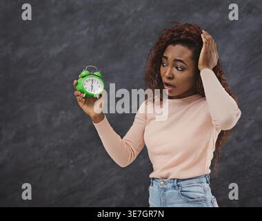 Femme noire bouleversée avec les cheveux bouclés blonds tenant et regardant le réveil debout avec la main sur la tête contre fond de tableau noir dans le studio Banque D'Images