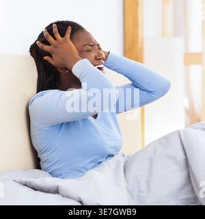 Femme noire avec des tresses portant un pyjama touchant la tête ayant mal à la tête assise sur le lit dans la chambre à coucher à la maison Banque D'Images