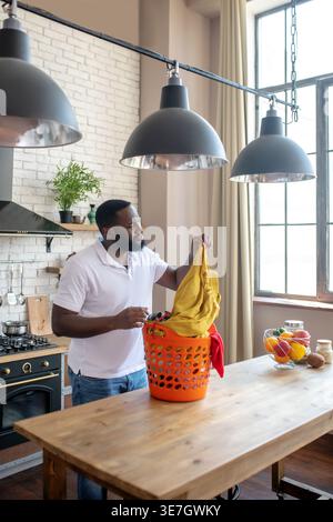 Homme noir malheureux prenant la chemise du panier tout en se tenant dans la cuisine Banque D'Images
