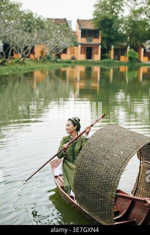 Femme asiatique dans des vêtements traditionnels regardant loin pagayer tout en étant assis sur le bateau dans l'étang pendant la journée Banque D'Images