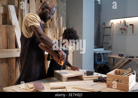 Fils noir dans un tablier portant des lunettes de protection perçant le bois debout avec père l'aidant de derrière dans l'atelier Banque D'Images