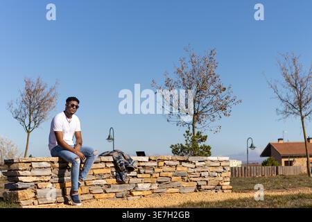 Homme noir dans des lunettes de soleil assis sur le banc de brique dans le parc le jour ensoleillé Banque D'Images