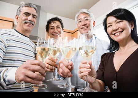 Groupe diversifié d'amis seniors grillant des verres à vin tout en se tenant debout dans la cuisine à la maison Banque D'Images