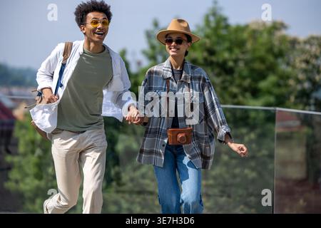 Couple de touristes noirs portant chapeau et lunettes de soleil tenant la main tout en courant près de la clôture de verre dans le parc le jour ensoleillé Banque D'Images