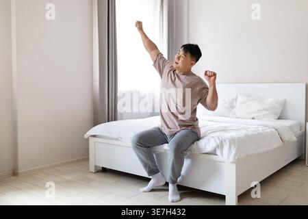 Homme asiatique étirant ses bras après s'être réveillé tout en étant assis sur le lit dans la chambre à coucher à la maison Banque D'Images