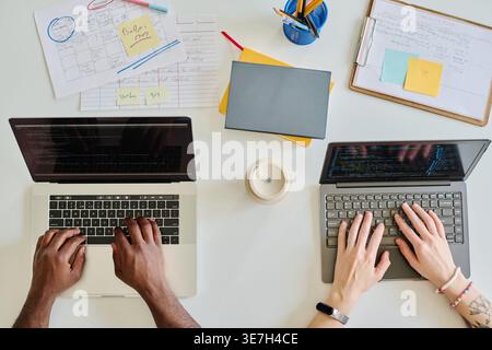 Vue à angle élevé de programmeurs noirs masculins et féminins caucasiens tapant des codes sur ordinateur portable tout en étant assis ensemble à table dans le bureau Banque D'Images
