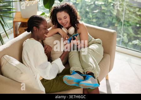 Femme mixte avec les jambes sur les genoux de colocataire femelle noire regardant la vidéo drôle sur les smartphones assis sur le canapé dans l'appartement pendant la journée Banque D'Images
