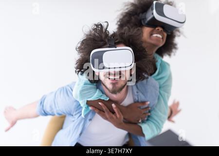 Couple noir et caucasien portant des casques VR ayant un plaisir piggyback équitation et expérimentant la réalité virtuelle sur fond blanc en studio Banque D'Images