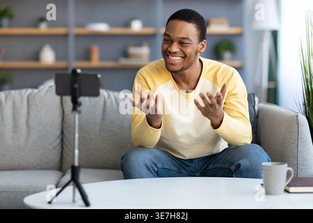 Homme noir souriant faisant du streaming en direct à l'aide d'un smartphone sur trépied donnant une leçon en ligne assis sur un canapé dans le salon à la maison Banque D'Images