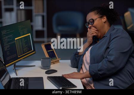 Femme d'affaires mixte fatiguée portant des lunettes couvrant la bouche tout en bâillant assis à table avec ordinateur et ordinateur portable travaillant tard dans la nuit en ... Banque D'Images