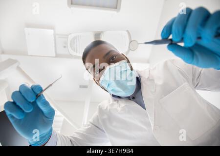 Dentiste masculin noir portant un masque facial et des gants tenant des outils dentaires prêts pour le contrôle en clinique Banque D'Images