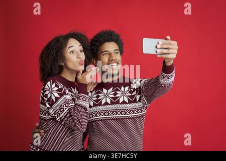 Couple noir avec afro portant un pull rouge soufflant baiser prenant selfie sur fond rouge en studio Banque D'Images