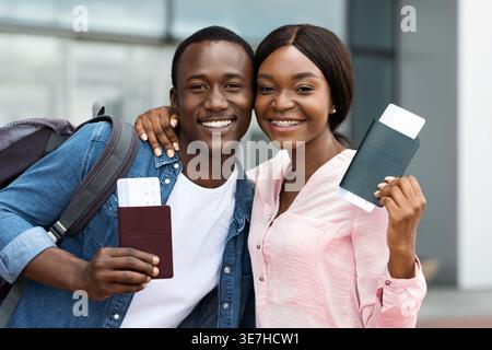 Couple noir souriant tenant des passeports avec billet d'avion debout près l'un de l'autre à l'extérieur de l'aéroport Banque D'Images