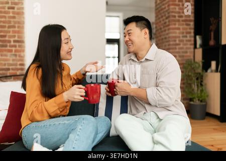 Couple asiatique s'amuser à boire du café et parler tout en étant assis ensemble sur le canapé dans le salon à la maison Banque D'Images