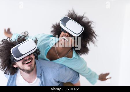 Couple noir et caucasien portant des casques VR ayant un plaisir piggyback équitation et expérimentant la réalité virtuelle sur fond blanc en studio Banque D'Images