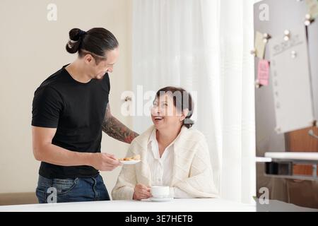 Fils métis souriant donnant à sa mère aînée assiette de biscuits tout en se tenant debout à table dans le salon à la maison Banque D'Images