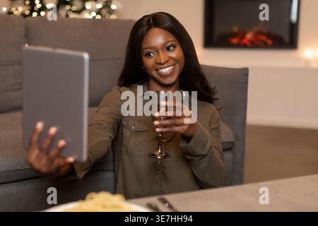 Femme noire ayant un appel vidéo sur tablette célébrant Noël tenant un verre de vin tout en étant assis dans le salon pendant le dîner à la maison Banque D'Images