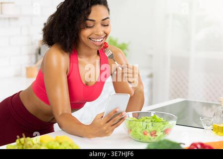 Femme noire portant un soutien-gorge de sport mangeant de la salade tout en utilisant un smartphone assis au comptoir dans la cuisine à la maison Banque D'Images