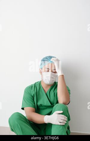 Chirurgien masculin asiatique stressé dans des maquillages verts touchant la tête assise sur le sol contre le mur dans le couloir de l'hôpital Banque D'Images
