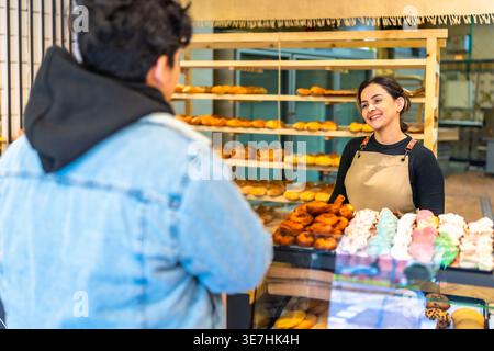 Boulangerie hispanique souriante dans le tablier parlant avec le client masculin achetant du pain tout en se tenant debout au comptoir avec différents pains dans la boulangerie Banque D'Images