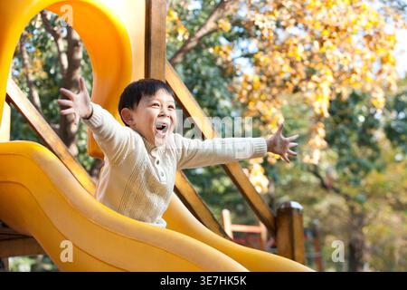Garçon chinois joyeux étirant les bras tout en étant assis sur le toboggan sur le terrain de jeu près des arbres d'automne pendant la journée Banque D'Images