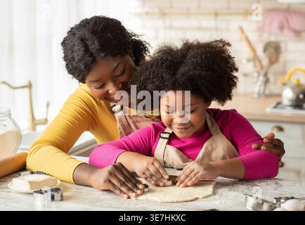 Mère noire et fille portant des tabliers cuisant des biscuits ensemble découpant différentes formes de biscuits de la pâte assise à table dans la cuisine à la maison Banque D'Images