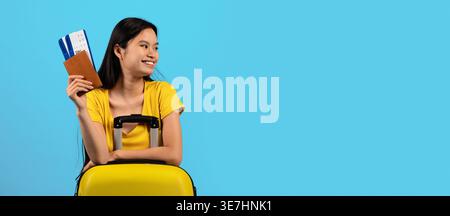 Adolescente asiatique souriante en chemise jaune avec valise et passeport avec billets regardant loin tout en se tenant debout sur fond bleu dans le studio Banque D'Images