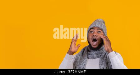 Homme noir choqué portant un bonnet tricoté et une écharpe ayant un appel téléphonique regardant loin debout sur fond jaune dans le studio Banque D'Images