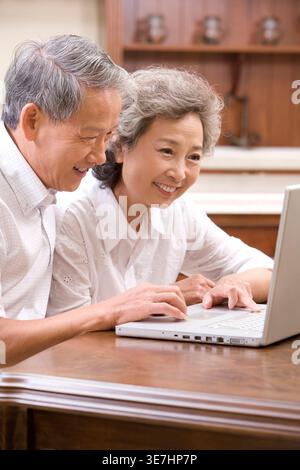 Couple chinois senior souriant utilisant un ordinateur portable tout en étant assis à table à la maison Banque D'Images