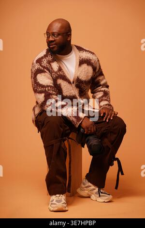 Photographe masculin noir portant des lunettes et un cardigan tenant la caméra regardant loin tout en étant assis sur une boîte en bois sur fond brun dans le studio Banque D'Images
