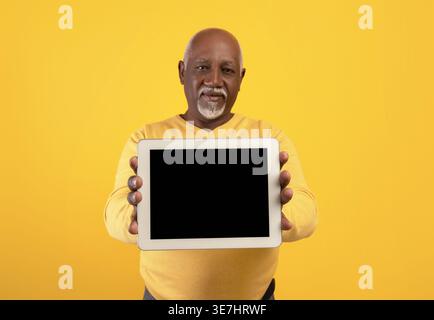Homme noir d'âge moyen tenant et montrant tablette avec écran blanc tout en se tenant debout sur fond jaune en studio Banque D'Images