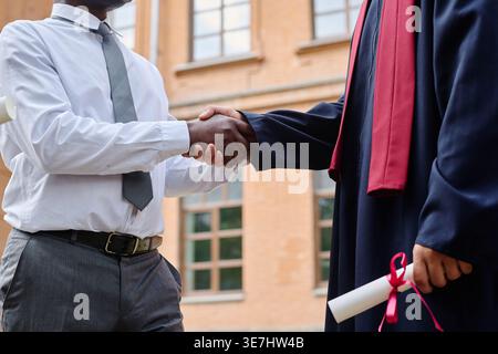 Professeur noir se serrant la main et félicitant l'étudiant masculin mixte après la cérémonie de remise des diplômes tout en se tenant sur le campus universitaire pendant da... Banque D'Images