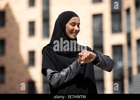 Femme musulmane du moyen-Orient souriante portant un hijab noir vérifiant son pouls sur le tracker de fitness debout dans la rue pendant la journée Banque D'Images