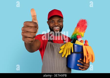 Chapeau de port et tablier noir nettoyant masculin montrant les pouces vers le haut et tenant le panier d'outils de nettoyage debout sur fond bleu dans le studio Banque D'Images
