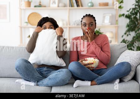Amies noires effrayées regardant un film ensemble tout en étant assis sur un canapé dans le salon à la maison Banque D'Images