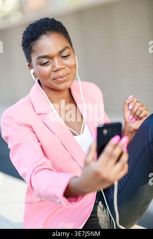 Souriant femme d'affaires noire en costume rose ayant un appel vidéo avec son smartphone assis à l'extérieur Banque D'Images