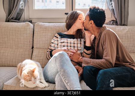 Homme noir et femme enceinte caucasienne embrassant assis sur le canapé avec chat à la maison Banque D'Images